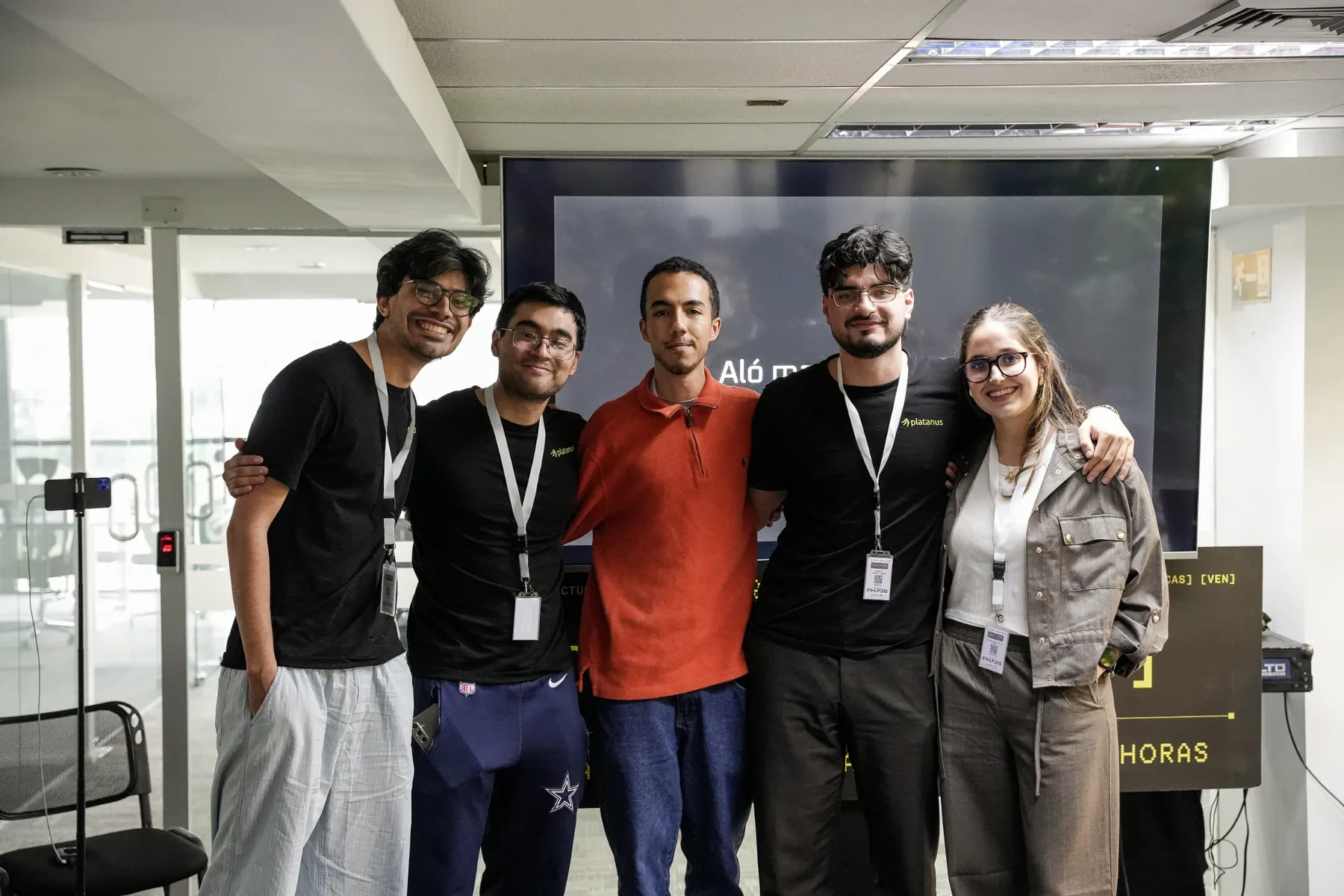 El equipo de Aló Mamá abrazado frente a la pantalla con la presentación, con las credenciales de Platanus Hack 26 al cuello